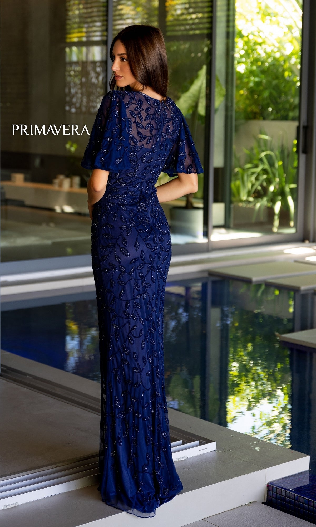A woman wearing the Long Prom Dress 13111 by Primavera in navy blue with a high neck and beaded details stands by an indoor pool, facing away, while large windows reveal lush greenery outside.